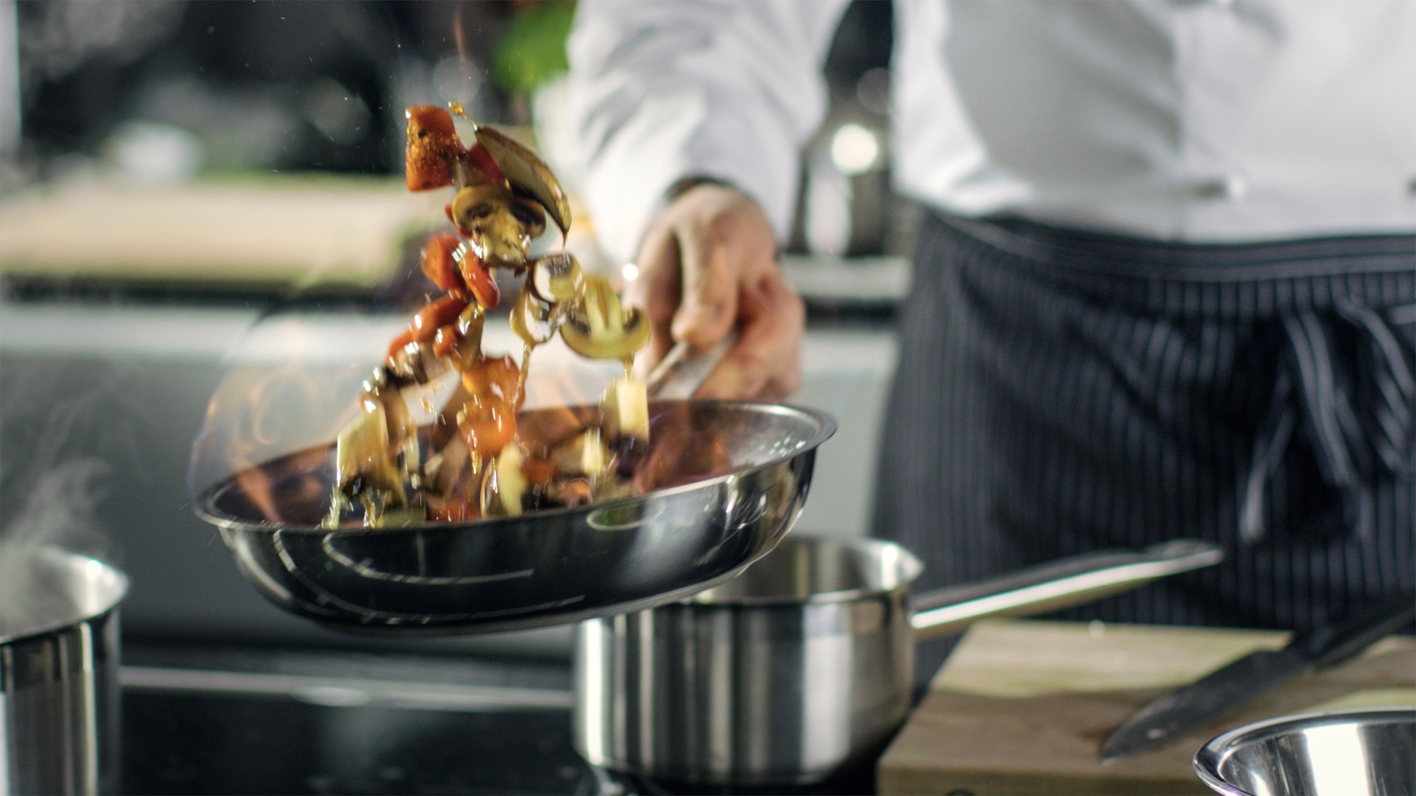Professional Chef Cooks Flambe Style. He Prepares Dish in a Pan with Open Flames. He Works in a Modern Kitchen with Different Ingredients Lying Around.